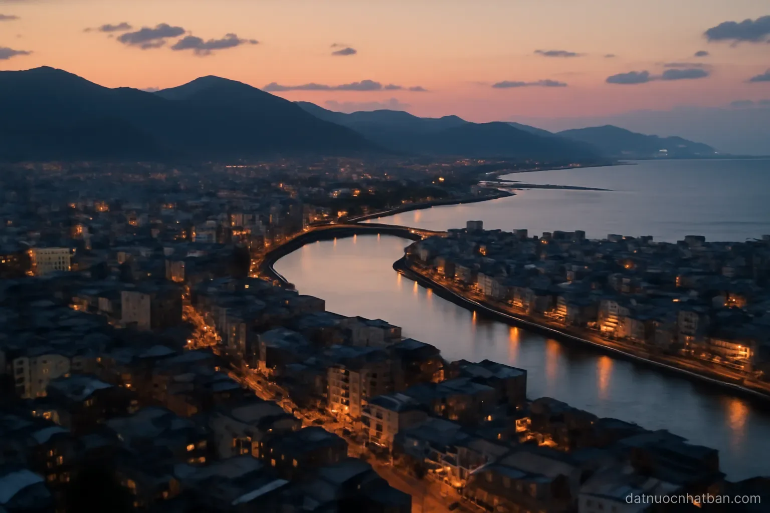 Aerial view of a Japanese coastal city at dusk with mountains behind and a river meeting the sea, warm lights beginning to appear
